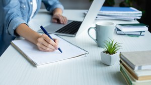 Foto eines Schreibtischs an dem eine Person sitzt. Die Person schreibt etwas auf einen Notizblock und hat gleichzeitig die andere Hand auf der Tastatur eines Notebooks liegen.