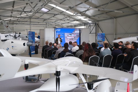 Teilnehmende einer Forschungsdemonstration in einem Flugzeughangar; im Vordergrund eine weiße VTOL-Drohne, im Hintergrund hält ein Referent einen Vortrag vor einer Leinwand.