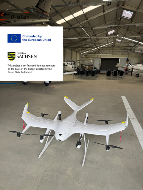 Weiße VTOL-Forschungsdrohne mit mehreren Rotoren und Tragflächen in einem Hangar mit mehreren bemannten Flugzeugen im Hintergrund.