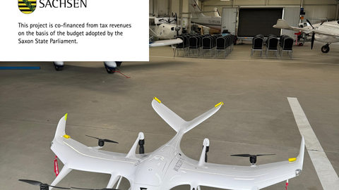 Weiße VTOL-Forschungsdrohne mit mehreren Rotoren und Tragflächen in einem Hangar mit mehreren bemannten Flugzeugen im Hintergrund.