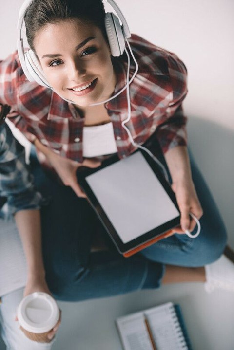 Das Foto zeigt eine junge Frau. Sie hält ein Tablet in der Hand und trägt Kopfhörer.