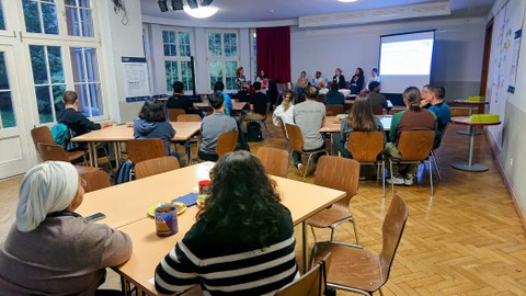 Foto von hinten in den Raum mit Blick auf Teilnehmer:innen und Panel