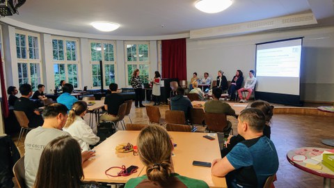 Foto von hinten in den Raum mit Blick auf Teilnehmer:innen und Panel