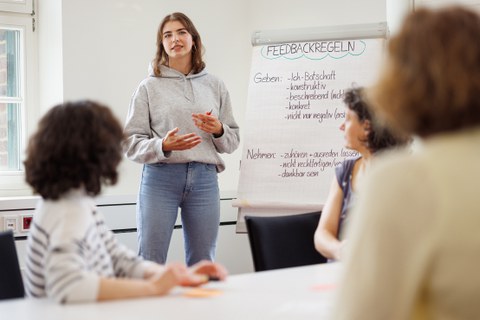 junge weibliche Person steht vor einer Flipchart mit Inahlten und erklärt etwas. Verwischt im Vordergrund sind Zuhörer:innen zu sehen.