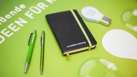 Bild eines grünen Tisches von oben. Auf dem Tisch liegt ein Notizbuch, grüne Kugelschreiber und ein Textmarker in Form einer Glühbirne. Alle Objekte tragen die Aufschrift "Career Service".