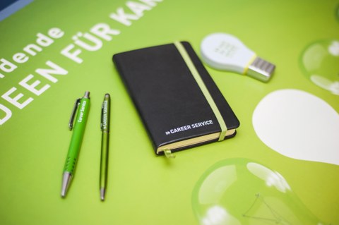 Picture of a table from above. On the table: Black notebook, pens, highlighter shaped like a light bulb. All objects marked with the inscription "Career Service"