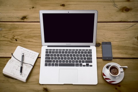 Laptop auf Schreibtisch, daneben ein Notizbuch, ein Smartphone und eine Kaffeetasse