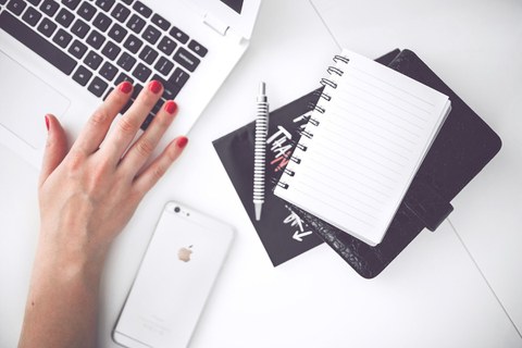 Laptop auf Schreibtisch, daneben ein Handy und ein Notizbuch. Eine Hand bedient den Laptop