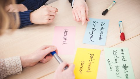 Auf einem Tisch liegen bunte beschriebene Karten. Man sieht die Hände von Personen, die an diesem Tisch sitzen und auf die verschiedenen Karten zeigen.