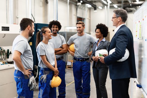 Gruppe junger Menschen in Arbeitskleidung mit Helmen in einer Fabrikhalle