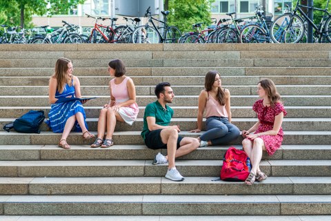 Foto: Fünf Studenten sitzen auf einer Treppe und unterhalten sich. Fahrräder stehen im Hintergrund. 