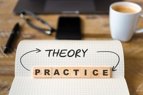 The photo shows a checkered block with the written word "theroy". The word "practice" is also depicted with wooden stones placed on top. The two words are connected by arrows. In the background you can see an office desk blurred.