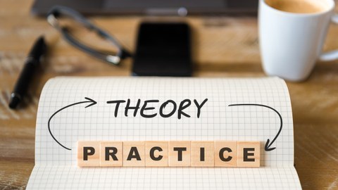 The photo shows a checkered block with the written word "theroy". The word "practice" is also depicted with wooden stones placed on top. The two words are connected by arrows. In the background you can see an office desk blurred.