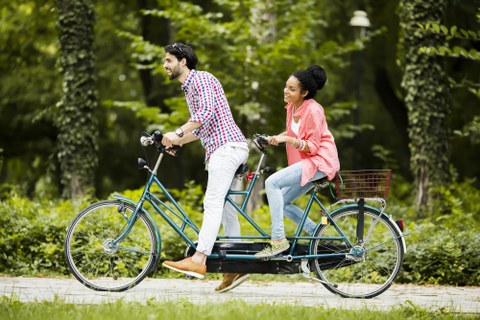 Das Foto zeigt zwei Personen. Sie fahren gerade mit einem Tandem durch einen Park. (Ein Tandem ist ein Fahrrad für zwei Personen.)