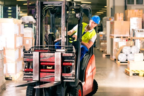 The photo shows a man with work clothes, helmet and yellow high-visibility vest in a warehouse. He is driving a red forklift truck.
