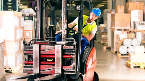 The photo shows a man with work clothes, helmet and yellow high-visibility vest in a warehouse. He is driving a red forklift truck.