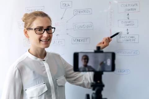 The photo shows a young woman in front of a whiteboard. She is speaking into her cell phone, which is filming the woman.