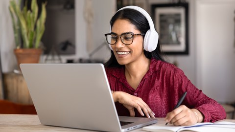 Smiling student woman wearing wireless headphones studying online sits at table at home using laptop and notebook