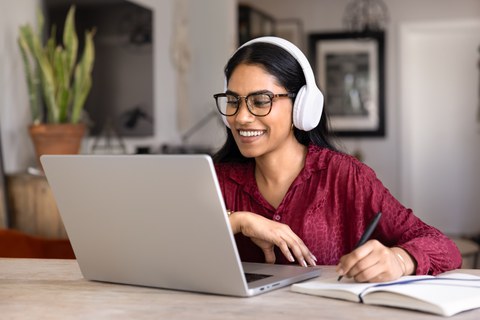 Lächelnde Studentin mit Bluetooth-Kopfhörern sitzt an einem Tisch vor einem Laptop und macht sich Notizen in einem Notizbuch
