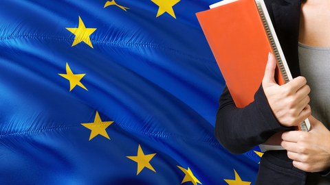  Junge Frau steht neben der der Flagge der EU. Hält ein oranges Buch mit Unterlagen.