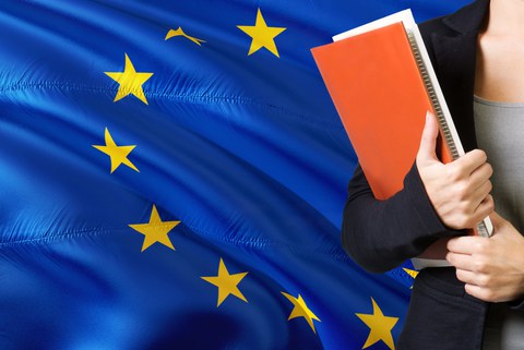  Junge Frau steht neben der der Flagge der EU. Hält ein oranges Buch mit Unterlagen.