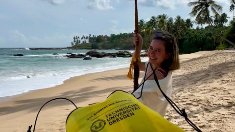Eine Person schaukelt am Strand; im Vordergrund hängt ein gelber Beutel der Technischen Universität Dresden.