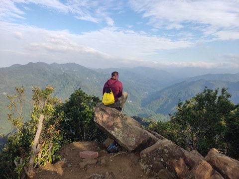 Eine Person sitzt auf einem Felsen am Rand eines Berges und blickt über eine weite, grüne Berglandschaft. Der Himmel ist leicht bewölkt.
