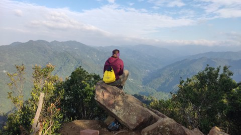 Eine Person sitzt auf einem Felsen am Rand eines Berges und blickt über eine weite, grüne Berglandschaft. Der Himmel ist leicht bewölkt.