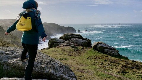 Eine Person mit gelbem Rucksack steht auf Felsen an einer windigen Küste und blickt aufs Meer.