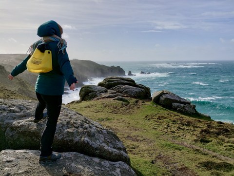 Eine Person mit gelbem Rucksack steht auf Felsen an einer windigen Küste und blickt aufs Meer.