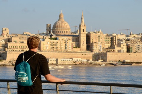 Eine Person steht an einer Uferpromenade und blickt über das Wasser auf eine historische Stadt mit großen Kuppeln und Türmen.