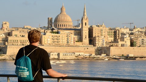 Eine Person steht an einer Uferpromenade und blickt über das Wasser auf eine historische Stadt mit großen Kuppeln und Türmen.