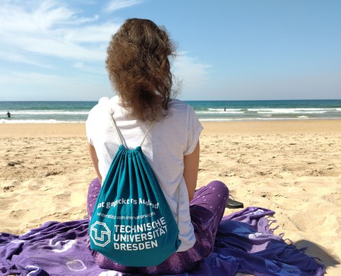 Eine Frau mit TUD-Turnbeutel sitzt auf einem hellen Sandstrand am Meer.
