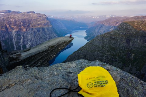 A student of TU Dresden sitting on Trolltunga, a famous rock jutting out high above a fjord and the surrounding mountains. In the foreground lies a TUD backpack.
