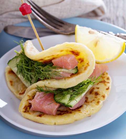 Dicker Pfannkuchen, der mit Lachs, Dill und Gurke gefüllt ist und von einem Spieß zusammengehalten wird. Auf dem Tellerrand liegt eine Zitronenscheibe.