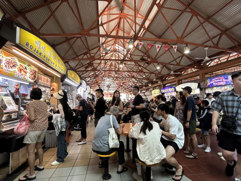 geschäftiger Food Court oder Hawker Centre: An den Seiten Essensstände, in der Mitte sitzen Menschen am Tisch