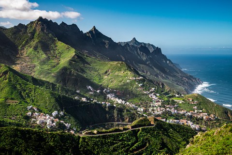 Auf einer Kanareninsel münden hohe, spitze Berge ins Meer. Sie sind leuchtend grün bewachsen und auf den niederen Hängen liegt ein weißgetünchtes Dorf.