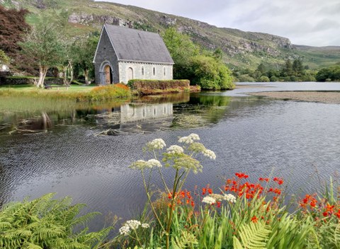  An old stone chapel standing on the shore of a lake. In the background, there is a rocky chain of hills, and in the foreground, marsh flowers bloom.