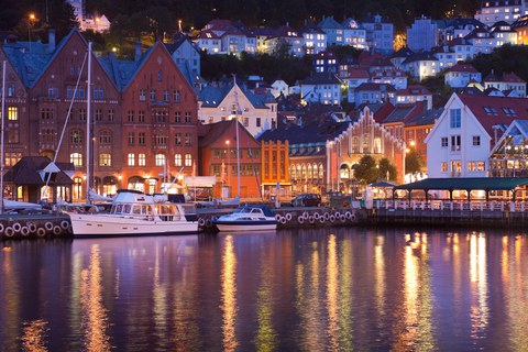 Bergen waterfront with a few boats and the facades of houses. Their evening window lights reflect on the surface of the water.