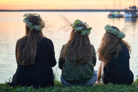 drei junge Frauen mit Kränzen aus Blumen und Kräutern sitzen am See