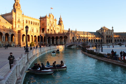 Menschen sitzen in Ruderbooten auf einem Kanal. Dahinter befindet sich ein langes halbkreisförmiges Gebäude mit einem Bogengang.