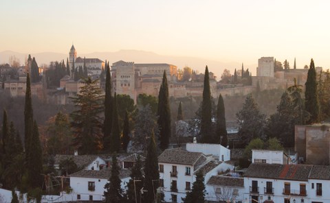 Hinter weißgetünchten Häuschen und Zypressen ragt eine große Burganlage aus rötlichem Stein auf. 