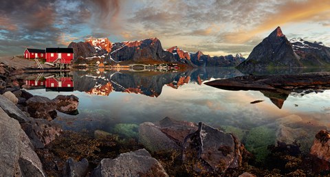 Wasser umgeben von schroffen, teilweise schneebedeckten Bergen in einer Dämmerungsstimmung. Am linken Rand befinden sich ein paar rote Häuser auf Stelzen.