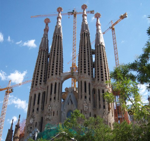 Eine Ansicht der Kathedrale Sagrada Familia mit vier hohen, schmalen Türmen voll dünner Fensterreihen, dahinter Baukräne.