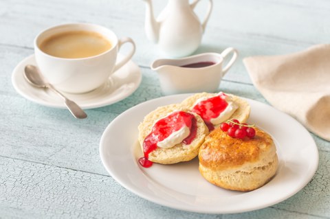 A plate of scones topped with clotted cream and jam. In the background, a cup of tea and a small teapot