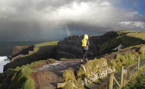 A person is walking along high cliffs covered with green vegetation. They are bathed in sunlight, and there is a rainbow above the cliffs.