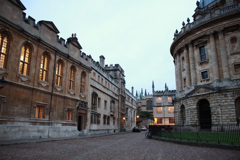 Oxford University Campus with fanciful old sandstone facades. Yellow light shines through the windows.