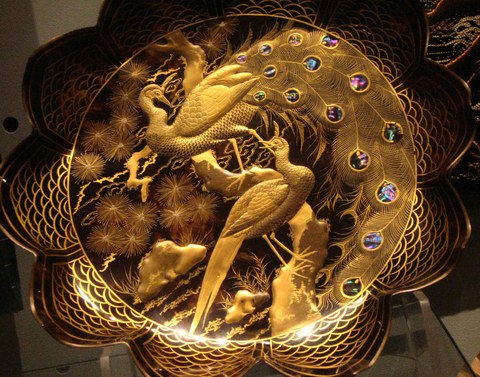 artistic plate of a dark material painted with golden peacocks in a Japanese-inspired style. Their tail feathers are decorated with mother of pearl