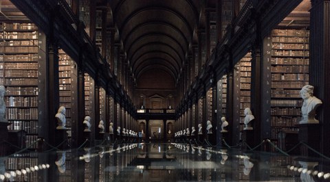 Blick auf einen Gang in einer alten Bibliothek. Zu beiden Seiten sind hohe Regale aus dunklem Holz. Der Gang in der Mitte ist gesäumt von weißen Statuen