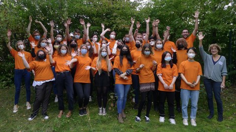 Eine Gruppe von Studierenden stehen zusammen vor einer Baumgruppe. Sie tragen orangefarbene T-Shirts, Mund-Nasen-Bedeckungen und strecken ihre Arme in die Höhe